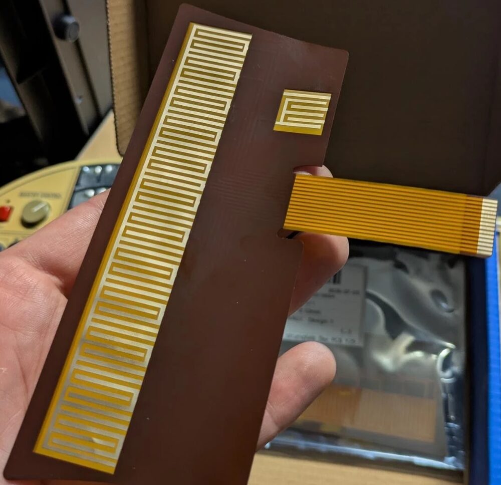 The new OM-27 strumplate flex PCB held in a hand: brown board with a long column of gold note contacts and comb, and an amber ribbon tail
