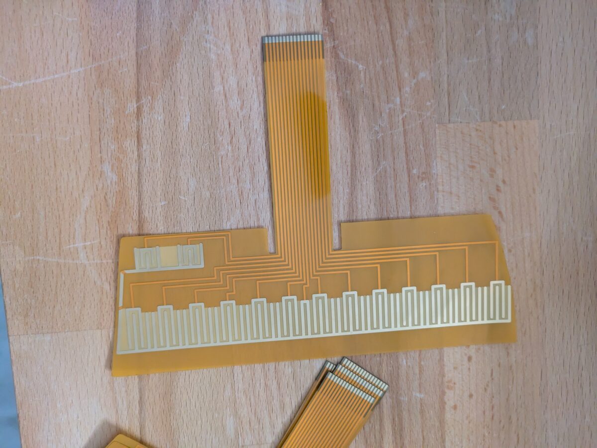 The new OM-84 strumplate flex PCB on the bench: amber film with the note contacts, the interleaved comb and the ribbon tail