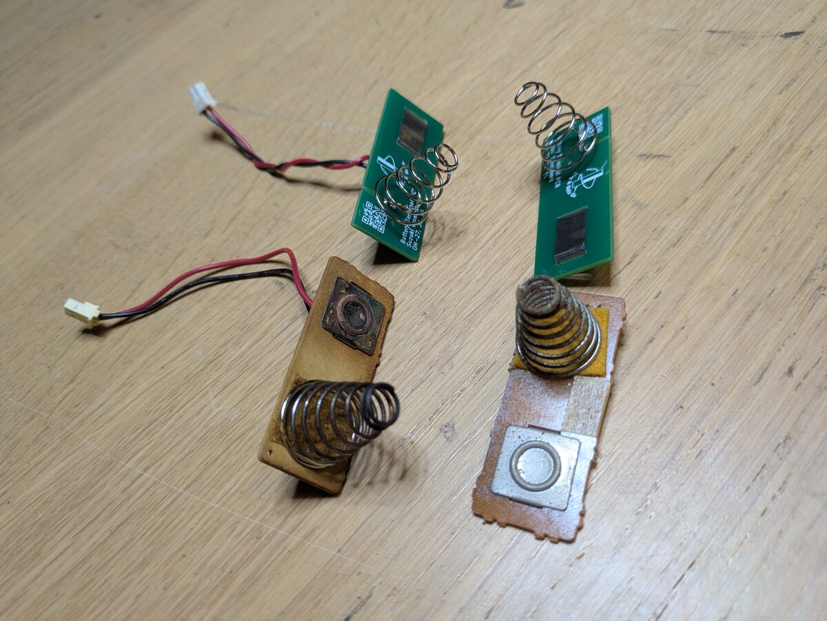 A new pair of green battery contact boards with springs, beside a corroded original pair